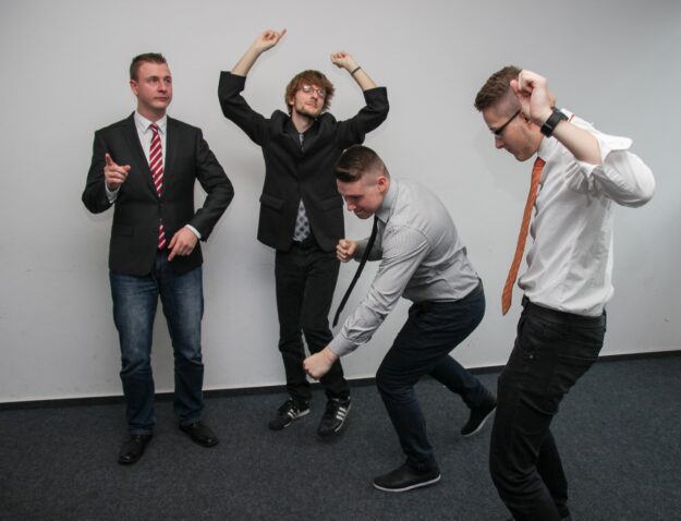 Four young businessmen playfully dancing, demonstrating adaptability and teamwork in a modern office setting. - Supply Chain Transparency