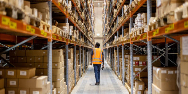 Warehouse worker walking down aisle of stacked boxes. Supply chain innovation. - Supply Chain Transparency